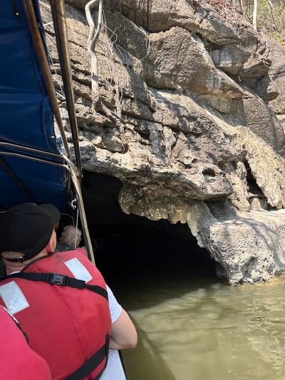 From a boat, a person in a red life jacket leans toward a rocky cliff face with a dark cave entrance. The cliff is textured and light-colored, contrasting with the dark opening. Water fills the cave's entrance. The boat has blue and metal railings. A person wearing a black baseball cap is visible. A rope and additional passengers are also present.