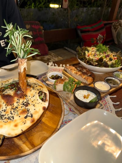 The image depicts an outdoor dining scene in what appears to be Baan Hin Kong, Thailand. A wooden table holds various dishes arranged for a meal. A large salad in a white bowl and several smaller bowls containing condiments or dips are visible. Bread, including naan-like flatbread sprinkled with black seeds, is also presented. One piece of bread has a sprig of rosemary protruding from it. A large white plate occupies the upper right of the frame, obscuring part of the scene. Pillows with Thai patterns sit on seats behind the table. The lighting suggests it's either late afternoon or evening. A person wearing a black shirt with white lettering is partially visible in the lower left corner.