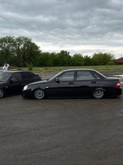 The image shows two black Lada Priora sedans parked on an asphalt surface. Trees line the background, with an industrial structure faintly visible behind them. The sky is overcast. A person in a white hooded garment stands near the first Lada. The location is potentially Pavlovskaya, Russia.