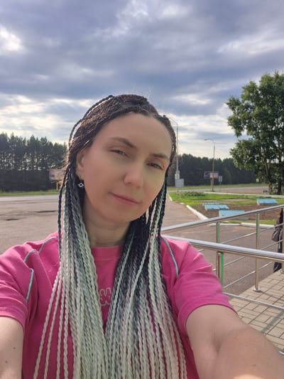 A medium shot captured outdoors shows a woman with long, braided hair of black, white, and gray hues. She is wearing a bright pink t-shirt. The woman is positioned in the foreground, with her face turned slightly towards the camera. The background features a road, a line of trees, and a cloudy sky. A metal railing is visible on the right side of the frame. The image appears to have been taken in the city of Mozhary, Russia.