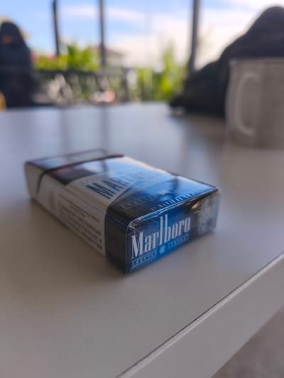 A closed pack of Marlboro cigarettes, with blue and white labeling, rests on a light-colored surface, likely a table. The pack is partially wrapped in clear plastic. In the blurred background, several out-of-focus figures are visible, along with some green foliage and a white mug. The overall setting appears to be outdoors, possibly a cafe or patio area. The location is identified as Фронтовики Снт, Russia.