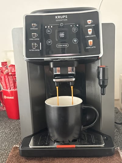 A Krups Sensation coffee machine is in operation. Coffee is being dispensed into a black mug. Beneath the machine, there is a red cup filled with EDAG-branded condiment packets. The scene is forensic in that the focus is on the coffee machine and its immediate surroundings. No people are visible. The location (Sanctorum, Mexico) is unspecified by any visual element in the photograph.