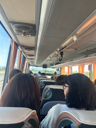 The image depicts the interior of a bus traveling along a highway. The perspective is from a passenger looking forward and slightly to the right. Several passengers are visible, mostly from the back or side of their heads. The bus interior features upholstered seats, overhead compartments, and ventilation grilles. Orange curtains are drawn over the windows, revealing a bright, sunny outdoor environment with some greenery and distant hills. The highway is visible through the windows, with other vehicles in the distance. The overall scene suggests a journey in a populated bus under clear weather conditions. The location of Polsko Padarevo, Bulgaria is not discernible from the visual information presented in the image.
