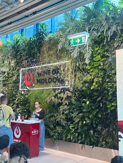 An event display featuring a vertical garden and a prominent sign for "WINE OF MOLDOVA" is pictured. The sign, illuminated by neon lights, is positioned in the center of the image, set against a lush backdrop of various green plants. Below the sign, a red display counter is set up, with bottles of wine and wine glasses arranged on its surface. A woman, likely an attendee or presenter, stands behind the counter, interacting with the display. A man wearing a yellow shirt and a backpack is visible to the left, observing the setup. The overall environment appears to be an indoor exhibition or event space, with a bright, modern ceiling structure and blue architectural elements visible in the background. The image is taken from a slightly elevated perspective, looking down towards the display.