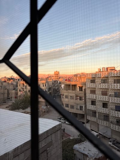 An elevated view captures an urban scene in Mu'adammiyat ash-Sham, Syria, during either sunrise or sunset, evidenced by the warm, low-angle light. The foreground is partially obscured by an out-of-focus black metal security grille and a fine mesh screen.
The midground displays a densely packed residential or mixed-use area composed primarily of multi-story buildings constructed from concrete blocks and light-colored plaster. Many structures exhibit signs of wear, unfinished sections, or ongoing construction, with exposed rebar visible on some rooftops. Numerous white and red water tanks are mounted on the flat rooftops. A prominent, taller building with a cylindrical water tower on its summit stands in the middle distance. A narrow street or alleyway winds through the buildings, accommodating several parked or moving white pickup trucks and a smaller white vehicle. Two individuals, clad in dark clothing, are visible walking on the street in the lower-middle portion of the image. A palm tree is situated near a building in the lower-left.
The background features a sky transitioning from pale blue at the zenith to vibrant orange and yellow near the horizon, where distant, light-colored mountains or hills are illuminated by the sun's low angle. A distinct, elongated, wispy cloud formation stretches horizontally across the sky.