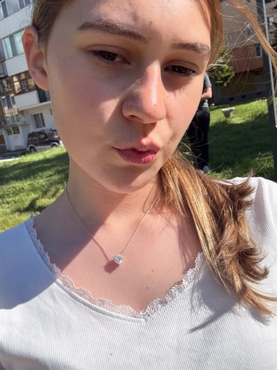 The image is a selfie taken by a fair-skinned individual with brown hair and eyes. They are wearing a white top with lace trim and a silver necklace with a heart-shaped pendant. The background shows a grassy area and a building. The building appears to be residential. Based on the background details, it is possibly taken in the city of Panagyurski kolonii, Bulgaria.