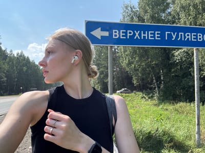 A young woman with blonde hair pulled back in a bun, wearing a black tank top and white earbuds, stands outdoors. She is looking to her left, her profile visible. A blue directional sign with white text and an arrow points to the left and reads "ВЕРХНЕЕ ГУЛЯЕВО" (Verkhnee Gulyaevo). Behind her, a grassy area with trees and a car on a slope is visible. The scene appears to be in Verkhnee Gulyaevo, Russia, indicated by the street sign.
