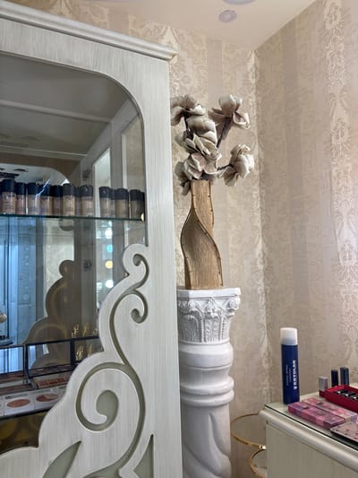 The image shows a close-up of decorative elements in what appears to be a well-lit indoor space, possibly a salon or a retail store, in Amman, Jordan. The primary subjects are decorative items and products.
Dominating the foreground is the top of an ornate white cabinet with carved scrollwork and an arched opening, revealing a mirrored interior displaying rows of glass bottles with black caps, likely makeup foundations. The cabinet itself is detailed with a distressed wood-grain finish.
Above the cabinet, a decorative arrangement features a twisted wooden element adorned with large, off-white fabric flowers. This is positioned next to a white, sculpted architectural element resembling a classical column fragment.
Further up, a shelf holds various makeup products. Prominently visible is a dark blue aerosol can labeled "KRYOLAN FIXING SPRAY." Next to it are several lipsticks and other cosmetic items arranged in a red organizer and transparent cases.
The background consists of wallpaper with a subtle damask pattern in light beige and cream tones. The lighting suggests it is daytime, with a warm and inviting atmosphere. There are no people visible in the frame, and the overall impression is one of tasteful display and product presentation. The location cues, such as the brand name on the spray and the general aesthetic, are consistent with a retail environment.