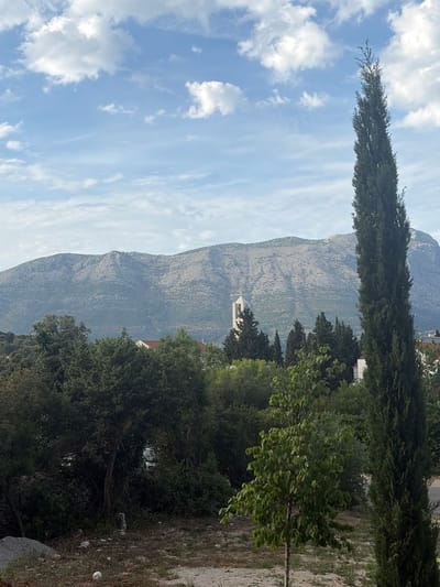 The image displays a panoramic landscape under a sky filled with scattered cumulus clouds against a light blue backdrop. In the background, a large, rugged mountain range, primarily grey and rocky with sparse vegetation, dominates the horizon. Below the mountain, slightly to the left of the frame's center, a tall, slender, light-colored church bell tower with a pointed roof and a small cross on top rises above the treeline. The tower features arched openings for bells. Adjacent to the tower and extending across the midground is a dense expanse of dark green trees and bushes, within which glimpses of white buildings with red tiled roofs are visible.
On the far right, a very tall, narrow, dark green cypress tree stands vertically, reaching almost to the top of the frame. In the lower-middle foreground, a smaller, lighter green deciduous tree with broad leaves is visible. The immediate foreground consists of dry, light-colored soil mixed with small stones and sparse patches of grass. A deflated white soccer ball rests on the ground in the lower-left corner, near some larger grey rocks and a low, partially obscured stone wall embedded within dense vegetation. No individuals or specific actions are depicted.
The scene is captured from a vantage point within the administrative city of Grad Korčula, Croatia, likely from an elevated position on Korčula Island. The prominent mountain range in the background is characteristic of the Pelješac peninsula, which is visible across the Pelješac Channel when looking north or northeast from Korčula Island. The vegetation and architectural elements are typical of the Dalmatian coastal region.