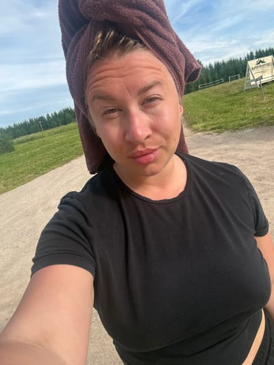A woman, with a brown towel wrapped around her head and blonde hair peeking out, takes a selfie outdoors. She is wearing a black t-shirt. The background shows a dirt road, green grass, trees, and a clear blue sky with some clouds. In the distance, there's a white sign with text, potentially in a Cyrillic alphabet, on an easel, and a small fence. The location is identified as Zamosh'e (Zaklinskoe S/P), Russia.