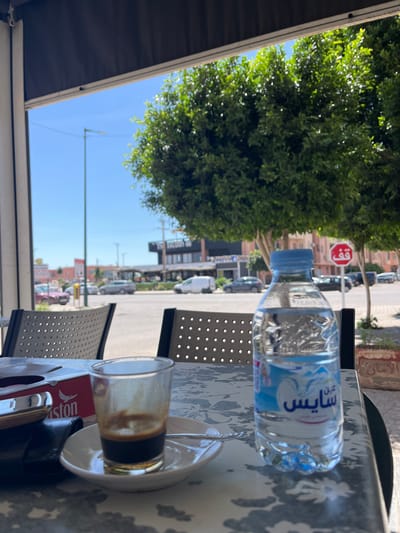 The image shows a table at an outdoor cafe in Saâda, Morocco. On the table is a partially consumed glass of coffee on a saucer, a bottle of "Saiss" brand water, and a red "Lipton" tea packet. In the background is a street scene featuring a tree, a stop sign in Arabic, buildings, and cars. The perspective is from the cafe, looking out towards the street.