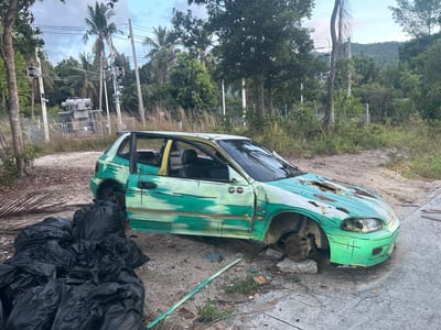 An abandoned, heavily damaged, green and white Honda Civic is on the side of a road in Baan Wang Ta Kien, Thailand. The car's paint is peeling, and rust is visible on the hood. The front passenger-side wheel is missing, exposing the brake assembly. The driver-side door is open. There are several large, black trash bags piled near the car. The background features lush tropical vegetation, including palm trees and power lines. A fenced-in electrical substation is visible in the distance. The sky is overcast.