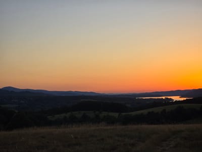 The image depicts a twilight landscape dominated by a gradient sky transitioning from pale blue at the top to vibrant orange and yellow near the horizon. Silhouetted against this colorful sky are undulating hills and mountains, covered in dark foliage. In the middle ground, a body of water, likely a lake or river, reflects the warm hues of the sunset. The foreground consists of dry, sun-bleached grass. The scene is devoid of human presence or activity, presenting a serene natural environment. The location is identified as Golyama Zhelyazna, Bulgaria.