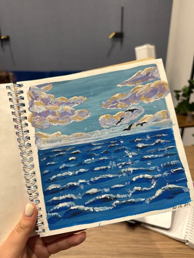A hand is holding a spiral-bound sketchbook open to a painting of a seascape. The painting depicts a blue sky filled with fluffy clouds in shades of white, yellow, and purple. Three black birds are flying across the sky. Below the sky is a choppy blue ocean with white-capped waves. The bottom right corner of the painting has a handwritten date: "20.02.24". The background of the image shows a blurred office environment with furniture and a plant. There is no information about Murom, Russia in the image.