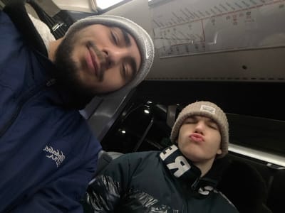 Two young men pose closely together in what appears to be a vehicle. The man on the left wears a blue jacket and a gray beanie, and he has a dark beard. The man on the right wears a light brown beanie and a dark green jacket with white lettering on the collar. A map is visible on the wall above them.