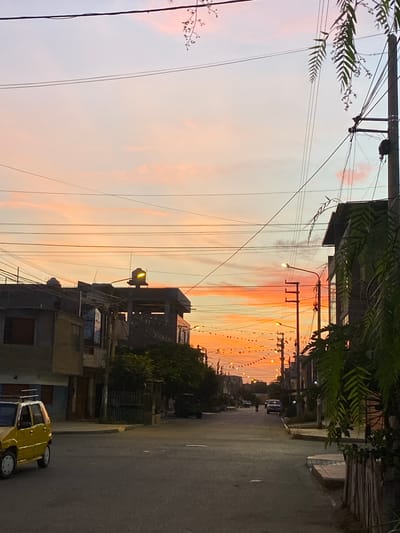 The image presents an eye-level street view during twilight in Chimbote, Peru. The sky transitions from light blue in the upper portion to vivid orange, red, and pink at the horizon, indicating a sunset with scattered clouds. Numerous dark utility lines span across the sky.
The paved asphalt street recedes into the background. On the left side, a yellow compact car with a silver roof rack is partially visible in the foreground, facing away from the viewer. Behind it, a series of multi-story concrete buildings line the street; one structure in the mid-left features a large, light-colored water tank on its roof reflecting the orange sky.
On the right, dark green, fern-like foliage hangs into the upper frame. A prominent dark utility pole with multiple power lines and associated equipment is situated on the far right. Further down the street, additional multi-story buildings and multiple illuminated streetlights are visible. A decorative string of small, triangular banners or lights is strung across the street overhead, extending into the distance.
Two indistinct human figures are walking on the right side of the street in the background. A light-colored sedan is also present further down the road on the right. A concrete sidewalk is visible on the immediate right. No specific actions or interactions beyond general movement are discernible. The scene depicts a residential or mixed-use urban environment.