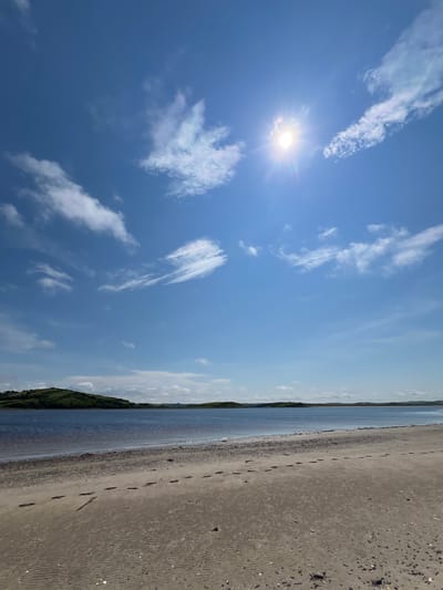 The image depicts a serene beach scene under a bright, sunny sky. The sun, a prominent radiant orb, is positioned in the upper right quadrant of the sky, casting a bright glare and lens flare. Wispy white clouds are scattered across the vast blue expanse.
The foreground of the image is a sandy beach with visible footprints leading away from the viewer. The sand displays rippled textures, suggesting the recent presence of water or wind. Scattered pebbles and small shells are also visible on the sand.
Beyond the beach lies a calm body of water, appearing as a deep blue. In the distance, a lush green, undulating landscape forms the horizon line. This landscape suggests rolling hills or a gentle coastline. The overall environment is natural and tranquil. The location is identified as Tullycullion, Ireland. There are no people or animals visible in the image.