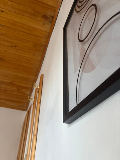 An upward-angled view captures an interior scene in the city of Antonovo, Bulgaria, depicting architectural elements and wall decor. The upper left portion of the image reveals a sloped ceiling constructed from horizontal light brown wooden planks exhibiting natural grain and some discoloration. Below the ceiling on the left, a light-colored wall features a segment of a wooden coat rack equipped with several brass-colored hooks. No items are suspended from the hooks. Dominating the right and central frame is a section of a white or off-white wall on which a rectangular framed piece of art is mounted. The art frame is dark, appearing black and matte, and slightly protrudes from the wall. The artwork within the frame displays an abstract design consisting of concentric and intersecting dark brown lines and circles, layered over soft, light brown or beige abstract shapes on a lighter background. Reflections are visible on the artwork's surface, indicating a glass or acrylic covering. No individuals or actions are present.