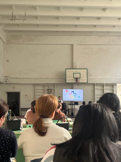 This media file depicts an indoor scene with several individuals present, seemingly attending an event or presentation. The setting appears to be a large, open space with concrete walls and tiered shelving units, reminiscent of a gymnasium or a community hall. A basketball hoop is visible mounted on one of the walls, suggesting a sports-related facility.

In the center of the image, a large screen displays a colorful logo featuring interlocking puzzle pieces, along with text that includes "genal" and other smaller words. The screen is positioned in front of a group of people, with some individuals facing the screen, suggesting they are the audience. The lighting suggests it is daytime, but the indoor environment makes it difficult to ascertain the specific time or weather conditions.

Several people are visible in the foreground and mid-ground, with their backs or profiles turned towards the camera. They appear to be dressed casually. One person in the mid-ground has distinctive blonde hair styled in pigtails and is wearing a white sweater. Another person in the foreground has long, dark hair. The overall mood seems attentive, with individuals engaged in watching the screen or listening to a presentation.

The presence of the basketball hoop and the open, multi-purpose space could indicate a community center or a school facility. The logo on the screen might be related to the organization or event taking place. No specific emotional expressions are clearly discernible due to the angles and focus of the people in the image. The location context of Gladzor, Armenia, is provided but not visually evident within the image itself.