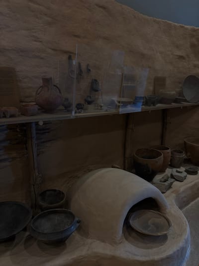 This image depicts an indoor museum exhibit located in Drama, Greece. The environment simulates an ancient dwelling or archaeological site, featuring textured, earth-toned walls.
The scene is divided into two main display areas. The upper section features a horizontal wooden shelf, supported by two vertical wooden posts. On the left side of the shelf, a partially visible informational sign in Greek text is affixed to the wall. The shelf displays various ceramic artifacts, including a prominent reddish-brown amphora-like pot with two handles, several small rectangular terracotta containers, and an assortment of smaller pots and fragments. Some of these items, along with hanging tools resembling ladles or spoons, are visible behind a clear glass display panel. A large, shallow, dark-colored bowl rests on the far right of the shelf.
The lower section of the exhibit showcases ground-level artifacts. Dominating the central-right foreground is a large, light-colored, domed clay oven, within which a shallow, dark plate or bowl is placed. Surrounding the oven on what appears to be an earthen floor are numerous dark and brown clay pots of varying sizes and shapes, some exhibiting visible cracks or wear. To the left foreground, two large, dark, shallow bowls or pans are present. Additionally, several grey, irregular stones, possibly grinding stones, are scattered near the pots and oven. The overall lighting is dim, emphasizing the textures and forms of the ancient artifacts. No animate subjects or actions are present.