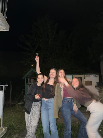 The image captures four young adult females outdoors at night in Tsar Kaloyan, Bulgaria. The background is dark, featuring silhouettes of trees and indistinct foliage, along with two small structures: a green wooden shed or playhouse visible to the left of the center, and a lighter-colored building with a dark opening further back on the right. A white vertical post or structure is partially visible on the far left.
From left to right:
1. A female, smiling, wears a dark top, a dark patterned open cardigan, and light-colored, vertical-striped, wide-leg trousers. She holds a short-stemmed glass containing a red liquid in her right hand.
2. A female, smiling, wears a black strapless top and blue flared jeans. Her right arm is raised, holding a champagne flute with a red liquid.
3. A female, smiling with an open mouth, wears a dark red top and blue flared jeans. An olive-green jacket is draped over her left shoulder and arm. She appears to be holding an object with her right hand, which is extended towards the person on the far right.
4. A female is in motion, captured with significant blur, leaning forward and smiling. She wears a dark brown long-sleeved top over a lighter undershirt and light-colored trousers. Her right arm is extended towards the third female, suggesting an interaction or gesture.
All subjects appear to be in good spirits, smiling or laughing, indicative of a social event or gathering. The scene is illuminated by an unseen artificial light source, highlighting the individuals against the dark nocturnal environment.