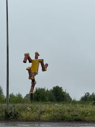 In Beloborodovo, Russia, an overcast sky provides diffuse lighting over a rural scene. A large, brightly colored sculpture resembling a robot stands prominently on a dark pole. The robot figure is constructed from what appear to be repurposed cylindrical materials, painted yellow and reddish-brown, with yellow circular elements at its joints. It is positioned in a dynamic, mid-stride pose. To the left of the sculpture is a tall, thin, dark pole. In the foreground, a low metal fence with a diamond pattern runs horizontally across the frame. Beyond the fence is a field of green grass interspersed with white wildflowers, leading to a line of dense green trees and shrubs that form the background. The ground in the immediate foreground is dark and appears wet, possibly muddy.