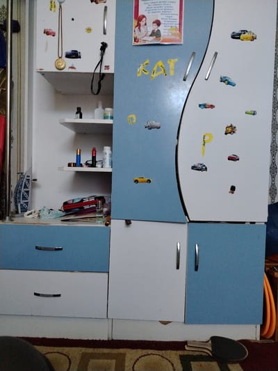 The image depicts a close-up, slightly angled, indoor shot of a cabinet unit with various items on display and attached to its surfaces. The cabinet has a light blue and white color scheme. The left section of the cabinet has shelves with items such as a gold pendant on a chain, a small black bottle, and other containers. Below the shelves are two drawers, one light blue and the other white, both with dark handles. The right section of the cabinet is predominantly light blue and is adorned with numerous stickers, primarily of cartoon cars, some appearing to be from the "Cars" franchise. There are also yellow letter stickers that spell out "KAT". A framed picture of two children studying with a teacher is also attached to this section. The cabinet unit is positioned in what appears to be a residential setting, with a patterned rug visible on the floor in the foreground. The presence of everyday objects and decorative elements suggests a personal living space. The image does not contain any visual cues that would allow for its location to be identified as Khiva, Uzbekistan.