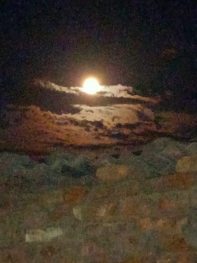 A full moon is visible in the night sky, partially obscured by clouds. The clouds are illuminated by the moonlight, creating a dramatic and somewhat abstract scene. The lower portion of the image appears to be a landscape, possibly trees or hills, with muted colors. There are no people or discernible structures visible in the image. The location is identified as Kyzyl Suu, Kyrgyzstan.