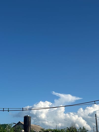 The image depicts a clear, bright blue sky on a sunny day, with a few fluffy white clouds visible on the right side. A power line runs diagonally across the frame from the top right to the bottom left, with a few small clips attached. Below the power line and clouds, there is a glimpse of a building with a grey, weathered roof and some green foliage. A black cylindrical object, possibly a water tank, is mounted on a structure next to the building. The overall scene suggests an outdoor setting, likely in a rural or suburban area given the presence of the power line and the building. The bright sky and distinct clouds indicate it is daytime, likely mid-morning or afternoon. There is no visible text in the image.