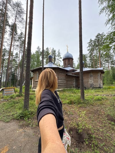 A selfie is taken in front of a wooden church with a blue dome and a golden cross in the village of Nenyamyaki, Russia. A woman with blonde hair is in the foreground, facing away from the camera and towards the church. She is wearing a black hoodie and dark pants. The church is surrounded by tall pine trees and green vegetation. The sky is overcast. To the left of the church, there is a small wooden structure.