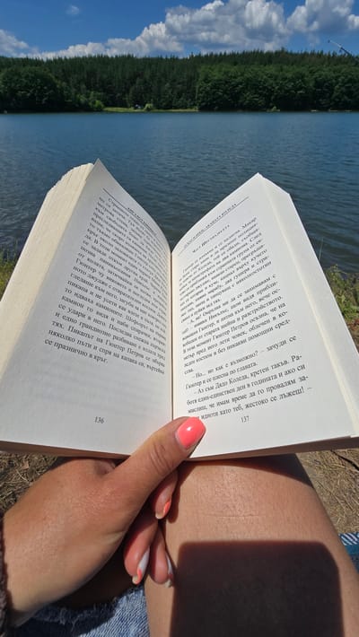 A first-person perspective shot shows a person holding an open book in front of a lake and forest. The book pages are visible, with text in Bulgarian and page numbers "136" and "137". The person's hand, with brightly colored nail polish, is holding the book. The background features a calm blue lake, a dense green forest, and a cloudy blue sky. The setting is likely outdoors, given the natural environment.