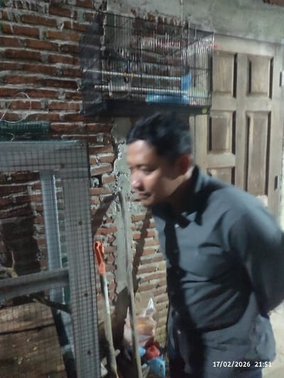 A person in a dark gray button-down shirt stands near a brick wall. Behind the person, a metal birdcage hangs on the wall, and a wooden door is visible to the right. To the left of the person, a metal cage is partially visible, along with a shovel and other tools. The photo appears to be taken indoors in a dimly lit area. The date and time "17/02/2026 21:51" are stamped at the bottom right corner of the image.