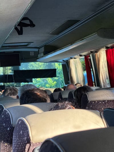 The image shows the interior of a bus, likely in the city of Kiosevtsi, Bulgaria, with passengers seated. The perspective is from a passenger looking forward. Overhead compartments and a strap are visible. Curtains, possibly red and beige, are drawn on the windows. The seats are upholstered, with some showing a dark pattern. The heads and upper bodies of several passengers are visible, their faces obscured by the angle and seating arrangement. Outside the windows, trees and foliage suggest a natural, possibly rural or suburban, environment. The lighting indicates it is daytime, with sunlight casting shadows inside the bus.