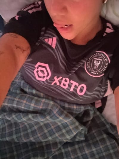 A person is lying down, wearing a black and pink soccer jersey with the words "XBTO" and "CLUB INTERNACIONAL DE FUTBOL" written on it. They are also wearing a plaid blanket, possibly in a hotel room. The person is looking directly at the camera, and their mouth is slightly open. The coordinates provided (latitude: -1.825, longitude: -80.752) correspond to a location in Montañita, Ecuador, specifically near the beach. It is not possible to determine the exact location within Montañita.