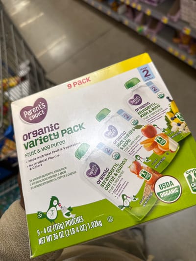 A slightly angled, close-up shot shows a box of "Parent's Choice Organic Variety Pack" baby food pouches being held in what appears to be a retail store aisle. The box, predominantly white and green, highlights its contents as "Fruit & Veg Puree."

The package is a "9 Pack" and states "NET WT 36 OZ (2 LB 4 OZ) 1.02kg," containing "9 - 4 OZ (113g) POUCHES." Key features advertised include "Made with Real Fruit & Vegetables," "No Artificial Flavors & Colors," and "BPA-Free Packaging." The "USDA Organic" seal is prominently displayed.

The variety pack contains three different organic purees, each in a "Stage 2" (likely 6+ months) pouch:
1.  Organic Blueberry, Kale & Rice
2.  Organic Root Veggies & Apple
3.  Organic Strawberry, Carrot & Quinoa

Each individual pouch also features the "Parent's Choice" brand and "USDA Organic" label. A small, stylized character with the text "GREAT FOR YOU" is visible on some of the pouch illustrations.

In the blurred background, typical retail shelving with various products and the handle of a shopping cart are visible, suggesting a supermarket or large retail chain in Worcester, United States. A person's arm or hand is partially visible in the bottom right corner, holding the box, indicating a shopping activity. The lighting suggests an indoor, daytime setting.