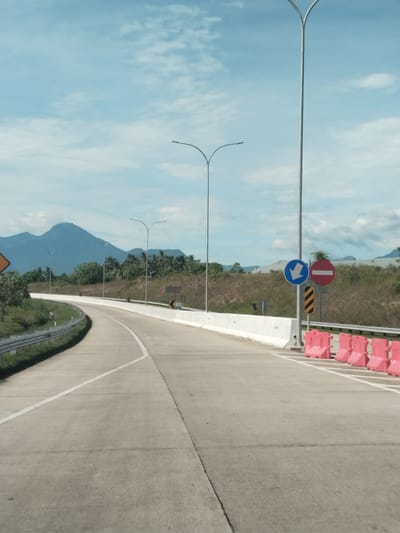 The image shows a paved road in Sihomlhok, Indonesia. The road curves to the left and is marked with white lines. On the right side of the road, there are pink barriers and a signpost with a blue arrow pointing down and a red circle with a horizontal line. Tall streetlights are visible along the road. In the background, there are mountains and trees under a cloudy sky. The scene appears to be taken during the day.