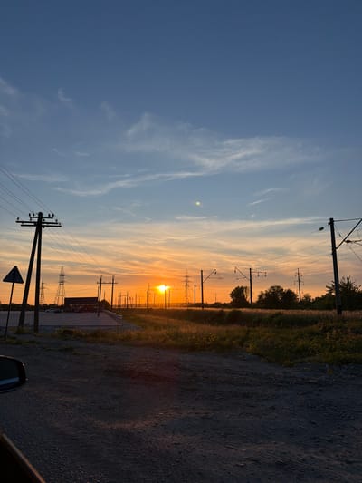 The photograph captures a rural landscape near the city of Yasenivtsi, Ukraine, during sunset. The sky displays a gradient of colors, transitioning from a deep blue at the top to vibrant orange and yellow hues near the horizon, with wisps of white clouds. Silhouetted against this colorful sky are numerous utility poles and electrical transmission towers, characteristic of power infrastructure. In the foreground, a dirt road with scattered gravel is visible, leading towards the background. To the left, the side mirror and part of a vehicle are in frame, suggesting the image was taken from a car. A triangular road sign stands to the left of the road. Further back, there is a small building with lights on, possibly a roadside establishment. Tall grasses and trees line the area beyond the road, also silhouetted by the setting sun. The overall impression is one of a quiet, end-of-day scene in a semi-rural setting with significant electrical infrastructure.