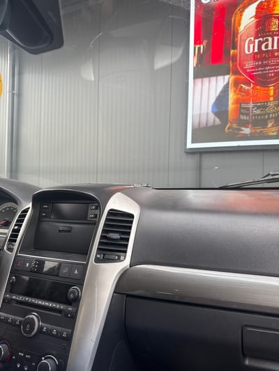 The image captures the interior of a car, focusing on the dashboard and windshield. A bright advertisement featuring a bottle of Grant's Triple Wood Blended Scotch Whisky is visible through the windshield. The dashboard is made of black plastic and features various controls, including an audio system and air vents. The environment outside the car appears to be a building with a ribbed metal wall. The scene does not provide any information that would allow for an identification of its specific location within Branipole, Bulgaria, nor does it depict any people.