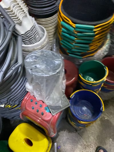 The image depicts an outdoor scene, likely a marketplace or storage area, in Ajuen, Indonesia. The ground is concrete and appears wet in places. The central focus is a collection of various plastic buckets and containers. In the foreground, a bright yellow lid-like object with a central hole is visible. To its left and slightly behind it is a red plastic container with a perforated pattern. To the right and behind the red container are several buckets, including a blue and yellow one, and a green and yellow one. Further back, more buckets are stacked, some with black bases and yellow and green banding. To the left of the image, a stack of white plastic items with a wavy pattern, possibly scoops or bucket handles, are visible, along with grey plastic items that appear to be part of a large container or structure. The overall impression is one of a commercial setting where plastic goods are being stored or sold. No people are clearly visible in the frame.