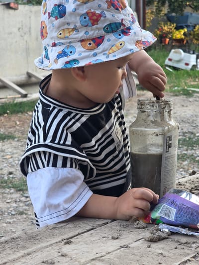 A young child, wearing a white bucket hat with a fish pattern and a black and white striped shirt, is intently engaged in an activity. The child's right hand is inside a glass jar containing a murky liquid and a stick, suggesting they are stirring or playing with the contents. To the right of the jar, a crumpled purple wrapper lies on a wooden surface. The environment appears to be outdoors, with a gravelly ground and hints of greenery in the background, along with a white toy vehicle. The image is taken from a medium close-up perspective, focusing on the child and their immediate surroundings. The overall scene suggests a moment of childhood exploration and play, possibly in a yard or a semi-rural setting.