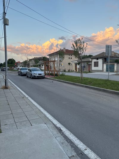The image depicts a quiet street scene in Popovo, Bulgaria, at dusk or dawn, indicated by the soft, warm light illuminating the cumuliform clouds in the sky with orange and pink hues.

In the foreground on the left, a concrete sidewalk composed of large square slabs shows minor signs of wear and sparse grass growing in cracks. A faded white line marks the transition from the sidewalk to the asphalt road. The asphalt road appears slightly reflective.

On the right side of the road, a silver SEAT Ibiza (Bulgarian license plate T 3477 AM, indicating Targovishte Province) is parked angled towards the curb. Behind it, a grey BMW sedan is parked parallel to the curb. A narrow grassy median strip separates the road from the adjacent structures. Within this strip, small, young trees are planted, and a wooden pergola structure with benches is visible, suggesting a public seating area.

Along the right side of the street, a sequence of buildings defines the background. The closest building on the right is an older, single-story structure with a tiled roof, white stucco walls, a brown window, and a prominent blue double-door entrance. Faint Cyrillic lettering is visible above the entrance. Adjacent to it stands a multi-story residential building with a light-colored facade, numerous windows, and a reddish-brown tiled roof featuring chimneys. Further down the street, additional buildings with similar architectural styles are visible.

On the far left, a utility pole rises vertically, supporting multiple power lines and a junction box. In the distance on the left side of the street, a "no entry" road sign (red circle with white bar) is visible, along with a yellow diamond-shaped "priority road" sign further back on the right. No people are visible in the scene.