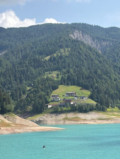 The image shows a vast expanse of turquoise water in the foreground, with a rocky, dry shoreline leading up to a steep, tree-covered mountainside. Several buildings are visible nestled on a grassy slope halfway up the mountain. The mountain range is densely forested with evergreens, and a rocky cliff face is apparent on the upper left side of the mountain. The sky above is partly cloudy with white clouds against a blue background. A black and white buoy is floating in the water. The scene depicts a serene natural landscape with human habitation integrated into the mountainside. The location is identified as Lateis / Latais, Italy.