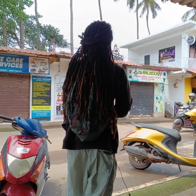 The image shows a person with long dreadlocks wearing a black hoodie with a floral design on the back and green pants, standing on a street in Oxel, India. In the background, there are buildings with signs for "We Care Services," "Insurance," "Overlockedd" (a clothing store), and "Urmila's Beauty Salon." Two motor scooters are parked on the street. A man wearing a helmet is riding one of the scooters.