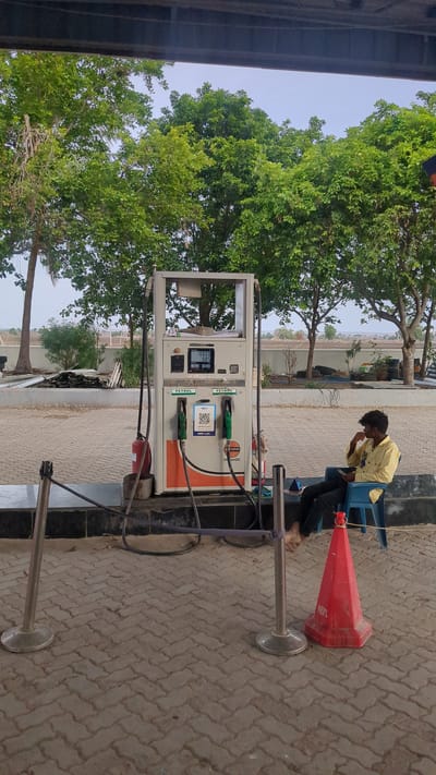This is a full-shot, eye-level, vertical, outdoor photograph of a gas pump at a gas station in Sugalimetta, India. In the center of the image is a gas pump with two nozzles. One nozzle is green, and the other is orange. The pump has a digital display showing "100.00 0.65" and other numbers. Below the display are QR codes for payment. The pump is branded "IndianOil". To the left of the pump is a red fire extinguisher mounted on a stand. A young man wearing a yellow shirt and dark pants is seated on a blue plastic chair to the right of the gas pump, looking at his phone. He is barefoot. A red traffic cone with "REPL" written on it is in the foreground to the right. Two metal stanchions with a rope tied between them are in the left foreground. The ground is paved with interlocking concrete blocks. In the background are several trees with green leaves, and a clear sky. The overall lighting is that of a bright, overcast day.