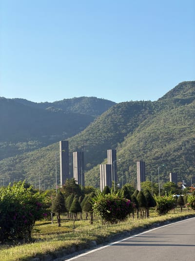 The image depicts a roadside view in Cam Lâm, Vietnam, characterized by a clear blue sky and lush green mountains in the background. In the mid-ground, several tall, narrow, dark grey, obelisk-like structures stand erected. In the foreground, a paved road curves to the right, bordered by a manicured grassy area with scattered conical trees and bushes laden with pink flowers. The scene is tranquil, with no people or animals visible, suggesting an early morning or late afternoon setting due to the shadows cast on the road. The overall environment appears to be a park or a landscaped area adjacent to a road, likely within a modern development or recreational zone in Cam Lâm.