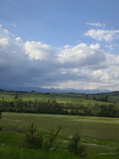The image depicts a landscape with a wide, green field in the foreground, leading to a line of trees. Beyond the trees is a rolling, green landscape with scattered trees and fields. In the distance, there is a mountain range with some snow-capped peaks. The sky is partly cloudy, with a mix of dark storm clouds and patches of blue sky. The location is likely near Dobrinishte, Bulgaria, given the mountainous backdrop, but it is impossible to confirm without ground truth. There is no indication of any people, subjects or interaction. It is difficult to determine any specific details about actions, interaction or objects.