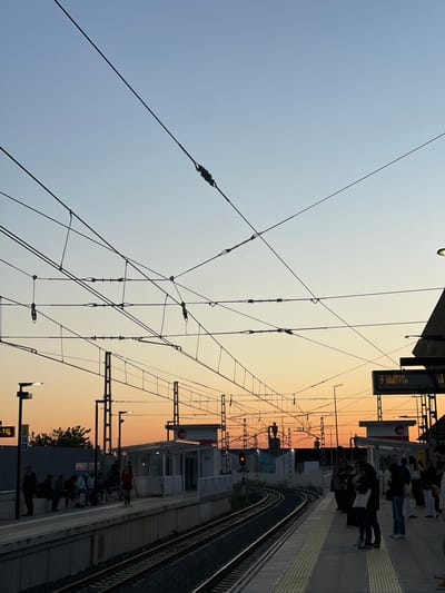 Here's a forensic description of the scene:

**Location:** A metro station in Paiporta, Spain.

**Environment:** A train station platform at dusk. The sky is a gradient from light blue to orange, indicating either sunrise or sunset. Overhead power lines crisscross the sky.

**People:** Several people are standing on the platform, seemingly waiting for a train. Most are facing towards the tracks.

**Objects:** The platform includes a train track, lights, signposts, and digital displays with train schedule information. The signs indicate that one destination is "Llíria" and "Maritim". Graffiti is visible on a wall next to the tracks.