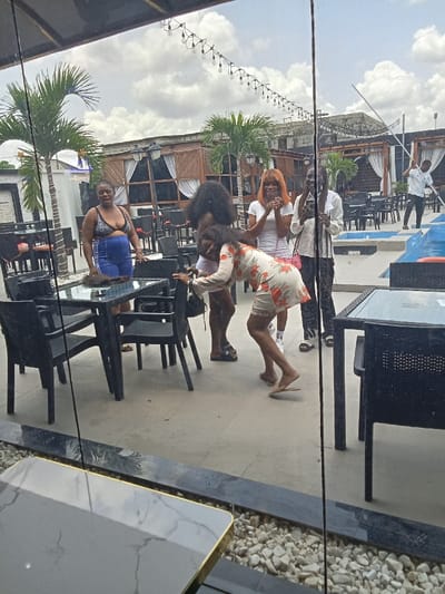 The image shows a group of five women and one man standing near a patio table with black chairs in an outdoor restaurant in Ujevwu, Nigeria. The women are wearing a variety of outfits, including a black bikini top and shorts, a white dress, a pink dress, and a white dress with a floral print. The man is wearing a blue shirt and dark pants. They are interacting with each other, and some are looking at the camera. The restaurant is surrounded by white rocks and palm trees. The outdoor area is also decorated with string lights. The scene appears to be a casual gathering at an outdoor restaurant in the city of Ujevwu. The location of the image is a restaurant, determined from the image's contents.
