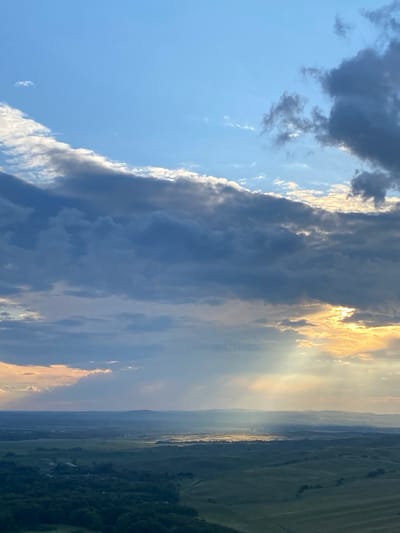 This is an aerial, wide-angle photograph of a rural landscape at sunset. The sky is a dominant feature, filled with dramatic, layered clouds. The upper portion of the sky is a clear, vibrant blue, transitioning to darker, textured gray clouds that stretch across the frame. Shafts of golden sunlight break through the clouds, illuminating patches of the landscape below. The middle ground features rolling hills covered in a patchwork of green fields and scattered trees. In the distance, a hazy horizon is visible, with faint outlines of more hills. The foreground shows a dense area of dark green trees. There are no people or specific structures clearly identifiable in the image, suggesting a natural, undeveloped setting. The scene evokes a sense of natural beauty and the grandeur of the sky at dusk.
