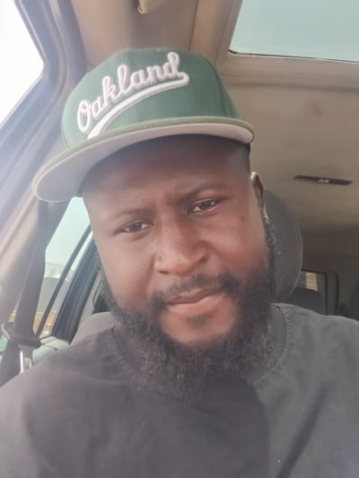 A man with a beard is wearing a green Oakland baseball cap inside a car. The location in Barakin Babele, Nigeria is not discernible from the provided image.